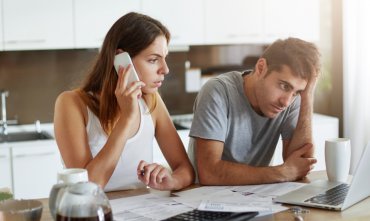 Couple in kitchen looking at bills and laptop while woman calls on smartphone