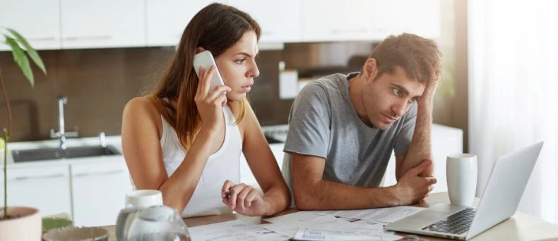 Couple in kitchen looking at bills and laptop while woman calls on smartphone