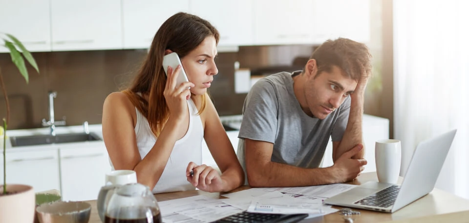 Couple in kitchen looking at bills and laptop while woman calls on smartphone