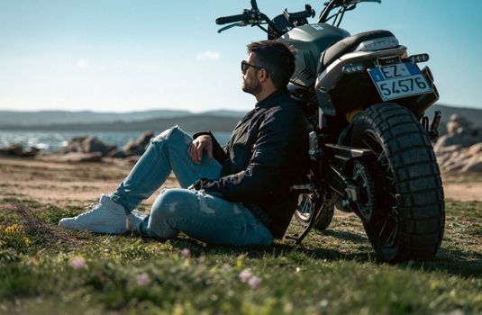 Man sits beside a parked motorcycle near the water, creating a calm riding moment for motorcycle insurance in Georgia.