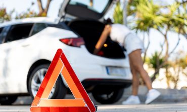 Red warning triangle on asphalt road with blurred woman checking car trunk in background, indicating car breakdown and road safety and the need for a car emergency kit.