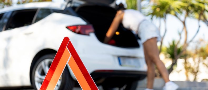 Red warning triangle on asphalt road with blurred woman checking car trunk in background, indicating car breakdown and road safety and the need for a car emergency kit.
