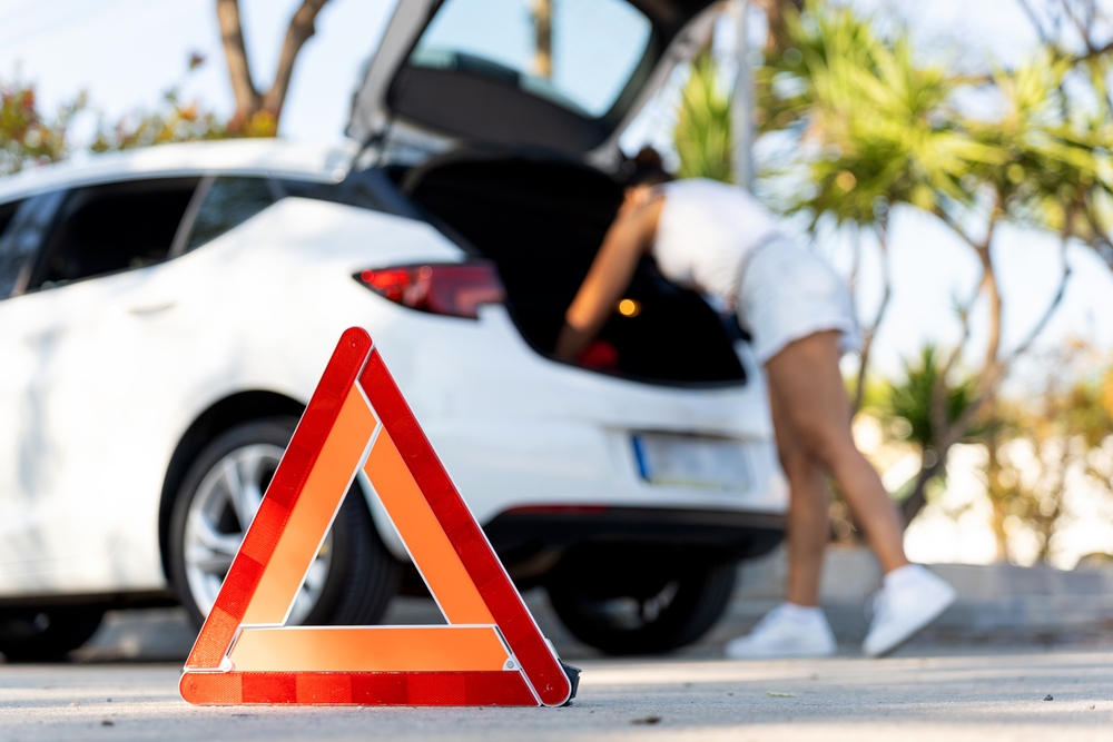 Red warning triangle on asphalt road with blurred woman checking car trunk in background, indicating car breakdown and road safety and the need for a car emergency kit.
