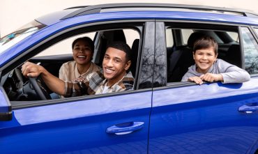 Cheerful parents and little son sitting in car together, happy spouses and male child looking out of window, ready for summer trip after they compared car insurance rates to find the lowest car insurance in Georgia.