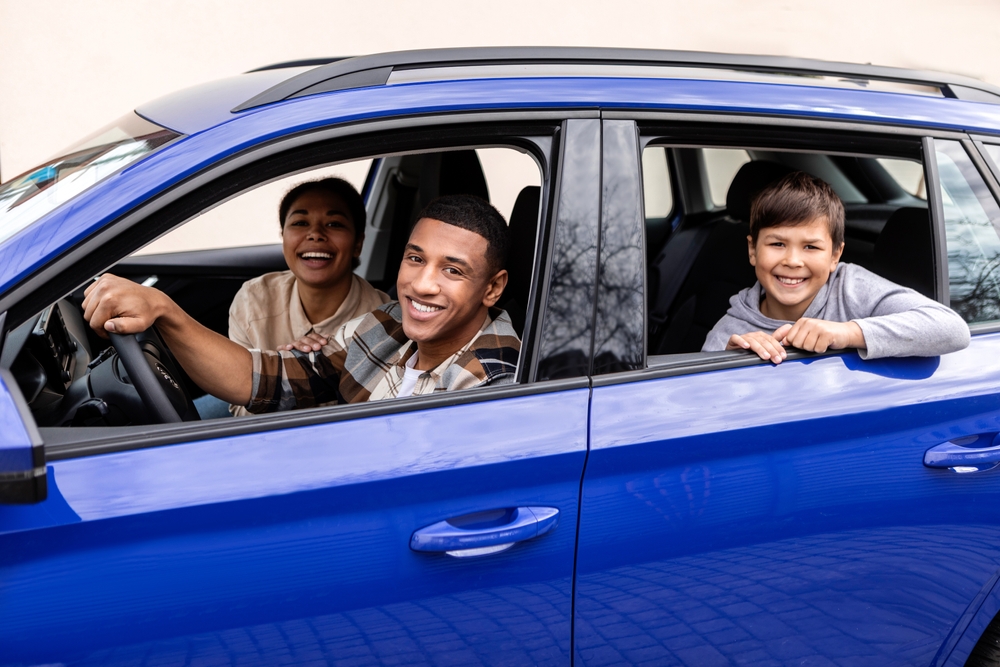 Cheerful parents and little son sitting in car together, happy spouses and male child looking out of window, ready for summer trip after they compared car insurance rates to find the lowest car insurance in Georgia.