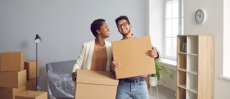 Two renters smiling while holding moving boxes in a new apartment, highlighting the importance of renters insurance to protect personal belongings during a move.