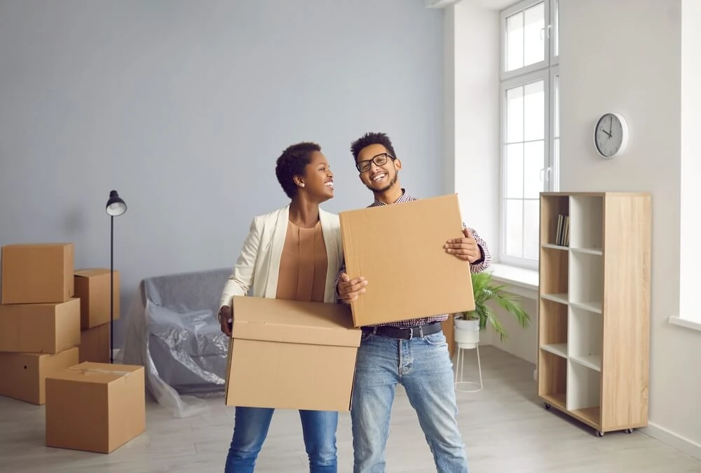Two renters smiling while holding moving boxes in a new apartment, highlighting the importance of renters insurance to protect personal belongings during a move.