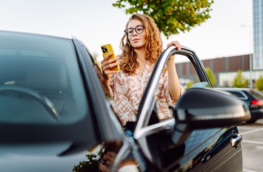 Woman standing by her car checking her phone, comparing options to switch car insurance quickly and conveniently.