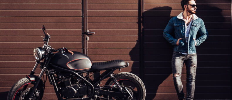 Handsome man stands against wall with his motorcycle parked next to him