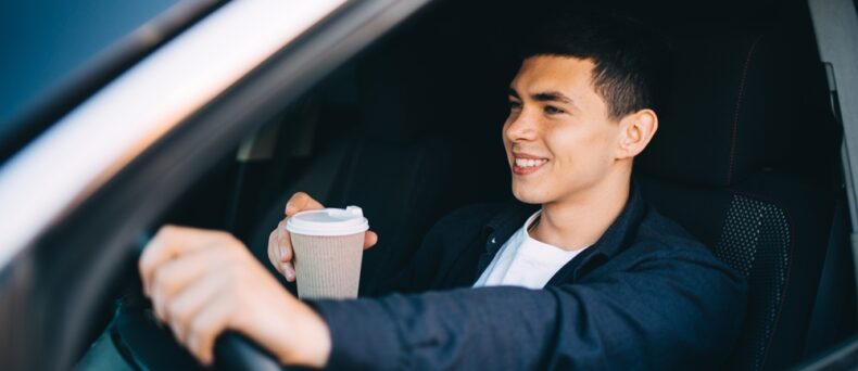 smiling young man behind the wheel with coffee cup
