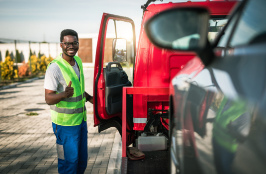 Smiling African American tow truck driver stands by open driver's door with car on back - cheap roadside assistance in Georgia
