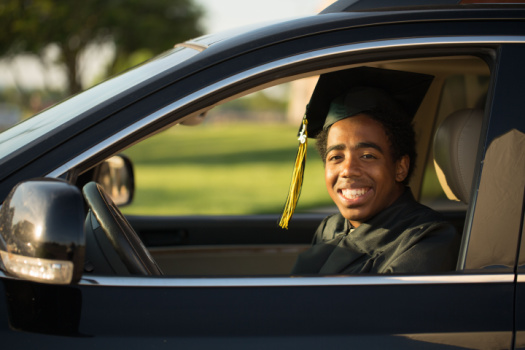 Smiling African-American graduate looks out his car window from behind the wheel - cheap car insurance for college students in Georgia.