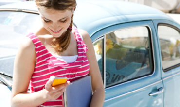 Young college student leans against her car while holding a notebook and texting - cheap car insurance for college students in Georgia.