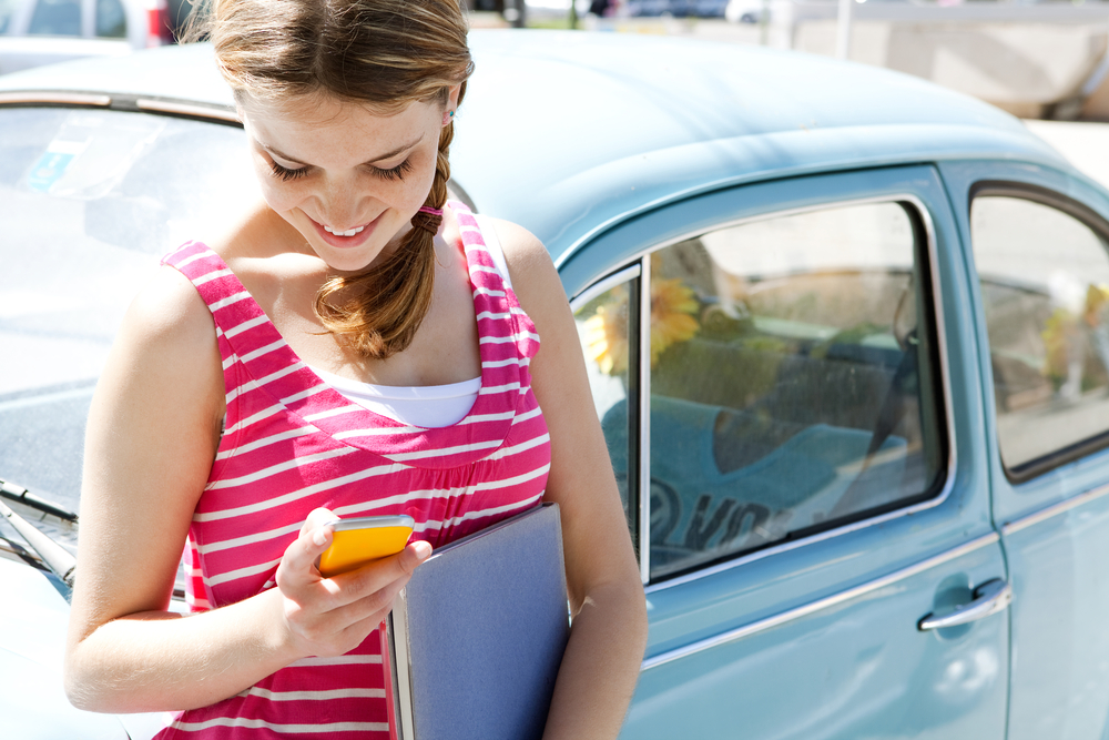 Young college student leans against her car while holding a notebook and texting - cheap car insurance for college students in Georgia.