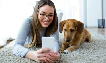 Young female on the floor in her apartment with her puppy looking at her phone.