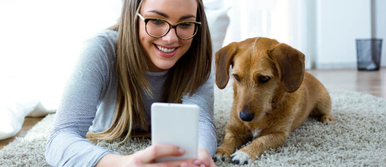 Young female on the floor in her apartment with her puppy looking at her phone.