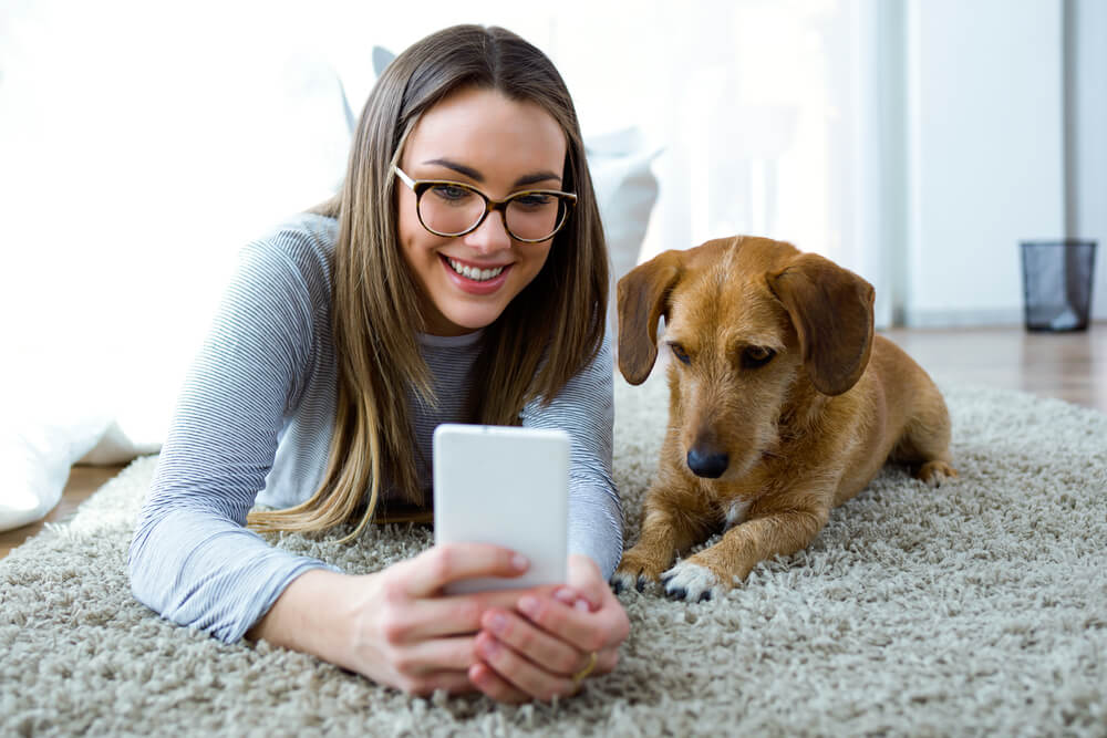Young female on the floor in her apartment with her puppy looking at her phone.