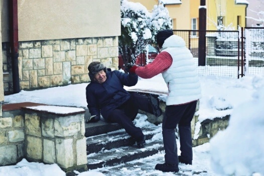 A person helps an elderly man up after he slips on icy, snowy steps.