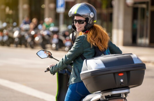 Smiling woman wearing a helmet sits on a motorcycle on a city street and looks back over her shoulder, highlighting questions like buy a motorcycle without a license.