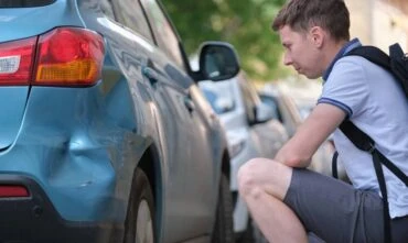 A man sits on the curb examining damage to his car from a hit and run driver - cheap car insurance in Georgia.