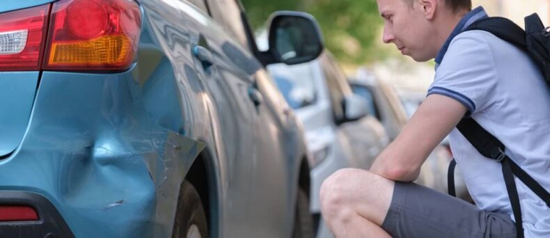 A man sits on the curb examining damage to his car from a hit and run driver - cheap car insurance in Georgia.