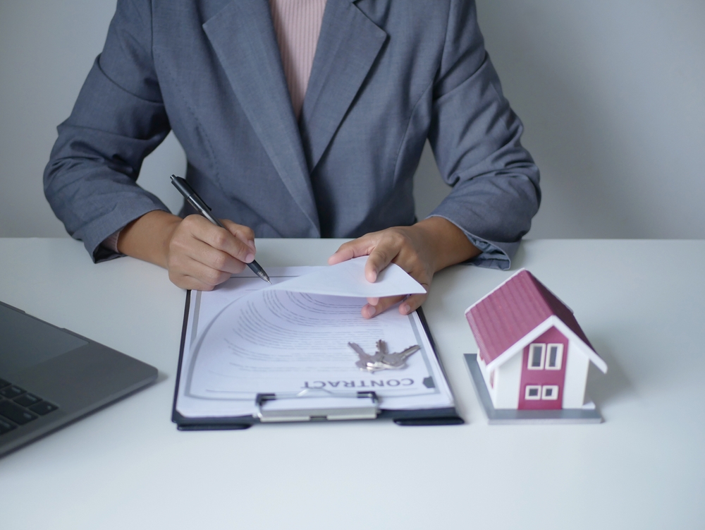 Person signing a housing insurance contract beside a model home, illustrating strategies for reducing renters insurance premium expenses through policy reviews, discounts, and better coverage choices.