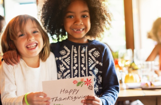 Two children smiling, holding a 'Happy Thanksgiving' card. Diverse kids, joyful moment, Thanksgiving celebration, friendship, happiness, festive atmosphere. Diverse kids holding Thanksgiving card.