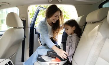 Mother buckling in her daughter in her car seat happy that she purchased the safest car of 2026.