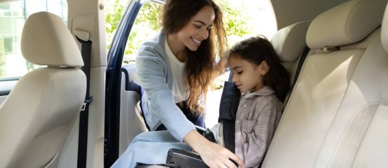 Mother buckling in her daughter in her car seat happy that she purchased the safest car of 2026.