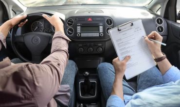 Defensive driving course in-car training session with a student driving while an instructor takes notes on a clipboard during a road skills evaluation.