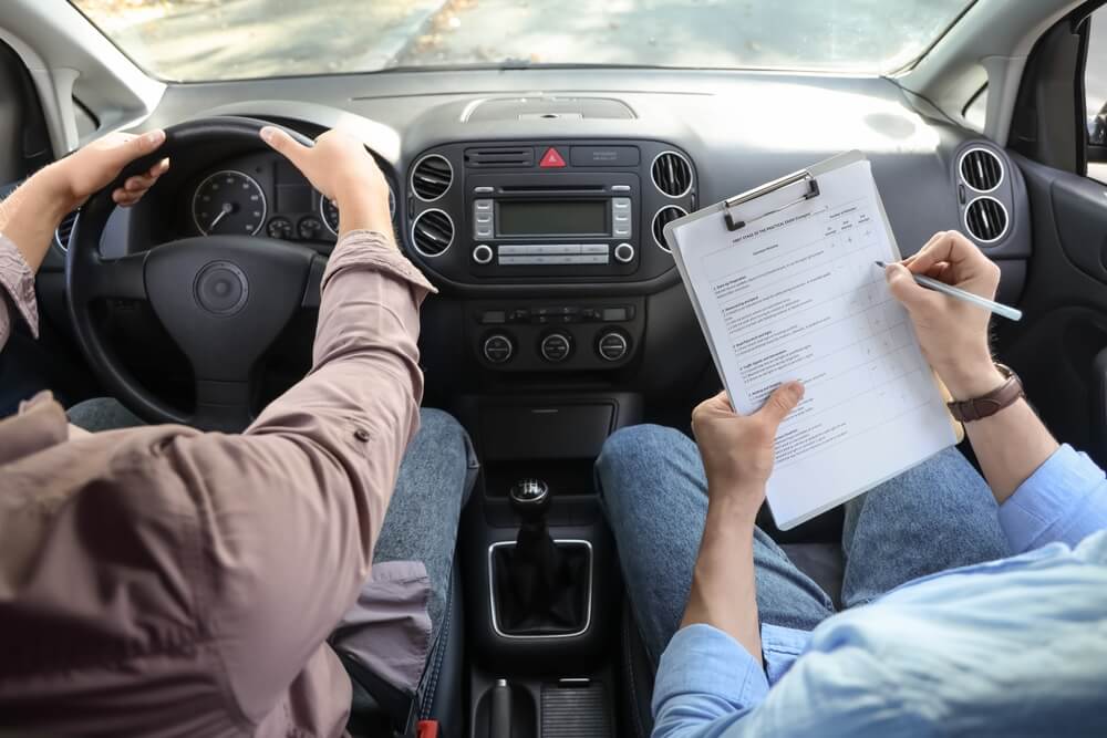 Defensive driving course in-car training session with a student driving while an instructor takes notes on a clipboard during a road skills evaluation.