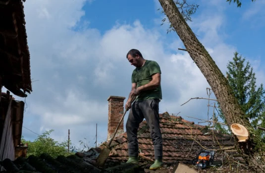 A man cleans the roof of a house after a big storm on a summer day. A man stands on the roof and removes fallen leaves and branches. Home insurance after a storm event.