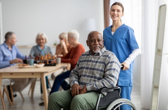 Happy black man older patient on wheelchair with female nurse smiling at camera, group of senior people sitting on couch on background, nursing home interior, healthcare for elderly people concept.