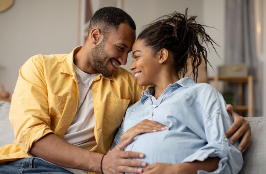 Pregnancy and childbirth and life insurance. Loving couple hugging expecting baby, husband touching pregnant wife's belly sitting on couch at home, when is a good time for a young family to buy life insurance.