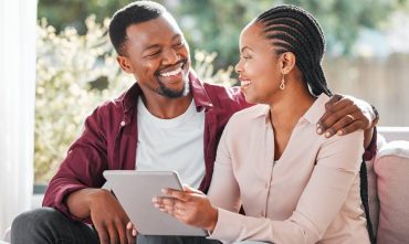 African American husband sitting on a sofa smiling and hugging his wife while she holds papers of her new policy after having used the open enrollment for life insurance.
