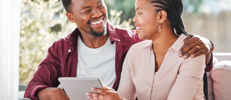 African American husband sitting on a sofa smiling and hugging his wife while she holds papers of her new policy after having used the open enrollment for life insurance.