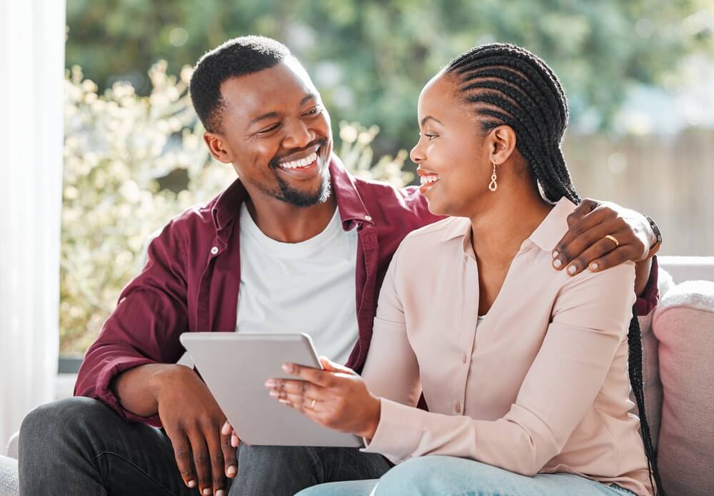African American husband sitting on a sofa smiling and hugging his wife while she holds papers of her new policy after having used the open enrollment for life insurance.
