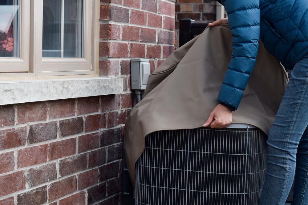 Woman putting cover on air conditioner unit outside the house during home fall maintenance checklist.