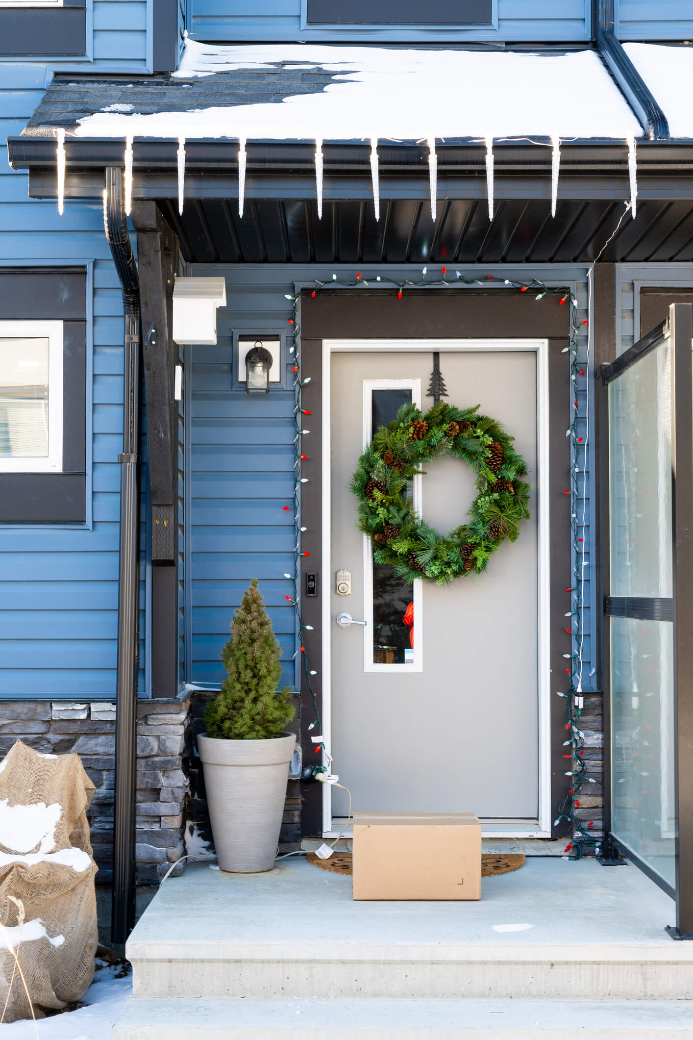 A package that has been delivered to a residential home in a suburban neighborhood is waiting to be picked up. It's winter and there is some snow on the ground. The house is decorated for Christmas and the holidays.