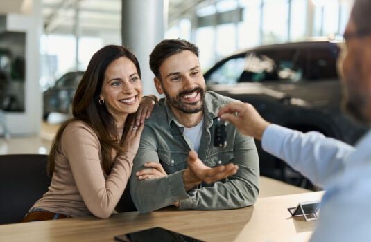 Happy couple receiving new car keys from a salesman during a meeting in showroom after learning the best time to buy a new car.