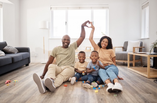 At home on the carpet, an African American family of four smiles and forms a hand-heart, symbolizing Georgia homeowners insurance.