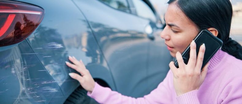 A woman examines scratches on the side of her car while speaking on the phone, documenting pre-existing car damage after a minor incident.