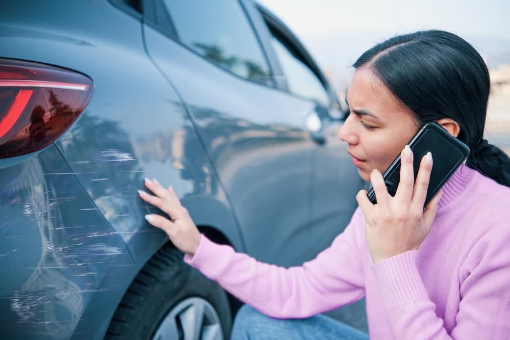 A woman examines scratches on the side of her car while speaking on the phone, documenting pre-existing car damage after a minor incident.