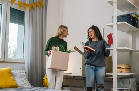 Two young women smile while unpacking boxes and books in a shared apartment, beginning independent living while balancing student loans renters insurance responsibilities.