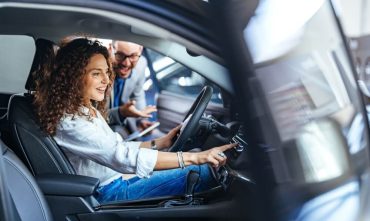 Curly-haired woman sits in the driver’s seat adjusting the dashboard controls while a salesperson leans in to explain features at a dealership, highlighting test drive car insurance.