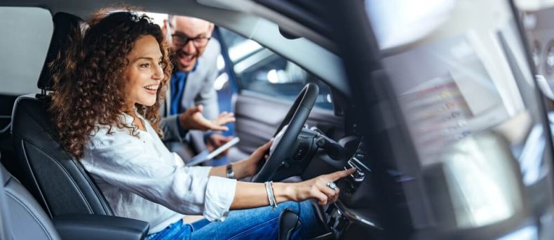 Curly-haired woman sits in the driver’s seat adjusting the dashboard controls while a salesperson leans in to explain features at a dealership, highlighting test drive car insurance.