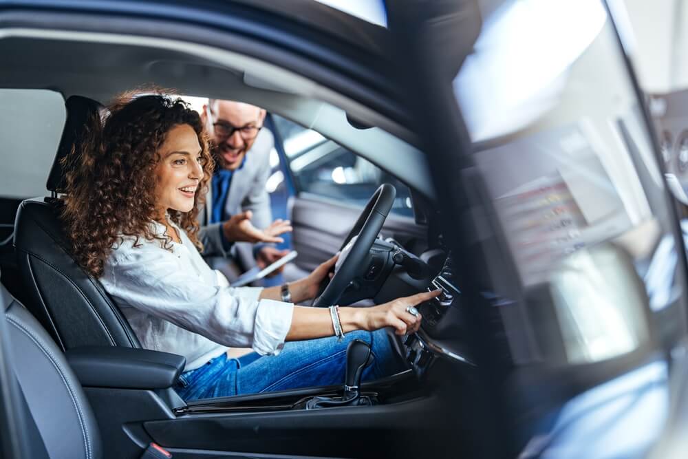 Curly-haired woman sits in the driver’s seat adjusting the dashboard controls while a salesperson leans in to explain features at a dealership, highlighting test drive car insurance.