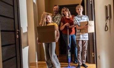 A smiling family of four standing in the hallway of a new apartment carrying moving boxes and a mop, representing the start of a new lease and the importance of choosing the right renters policy deductibles.