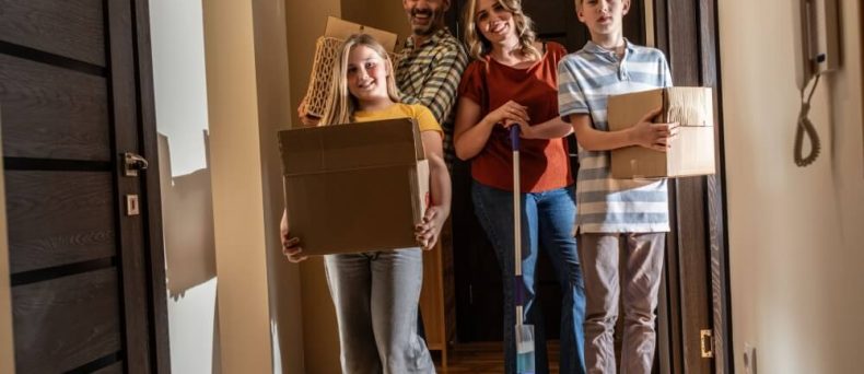A smiling family of four standing in the hallway of a new apartment carrying moving boxes and a mop, representing the start of a new lease and the importance of choosing the right renters policy deductibles.