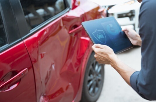 Person inspecting damage on a red car with a tablet, representing how no down payment car insurance can help cover accident costs quickly.
